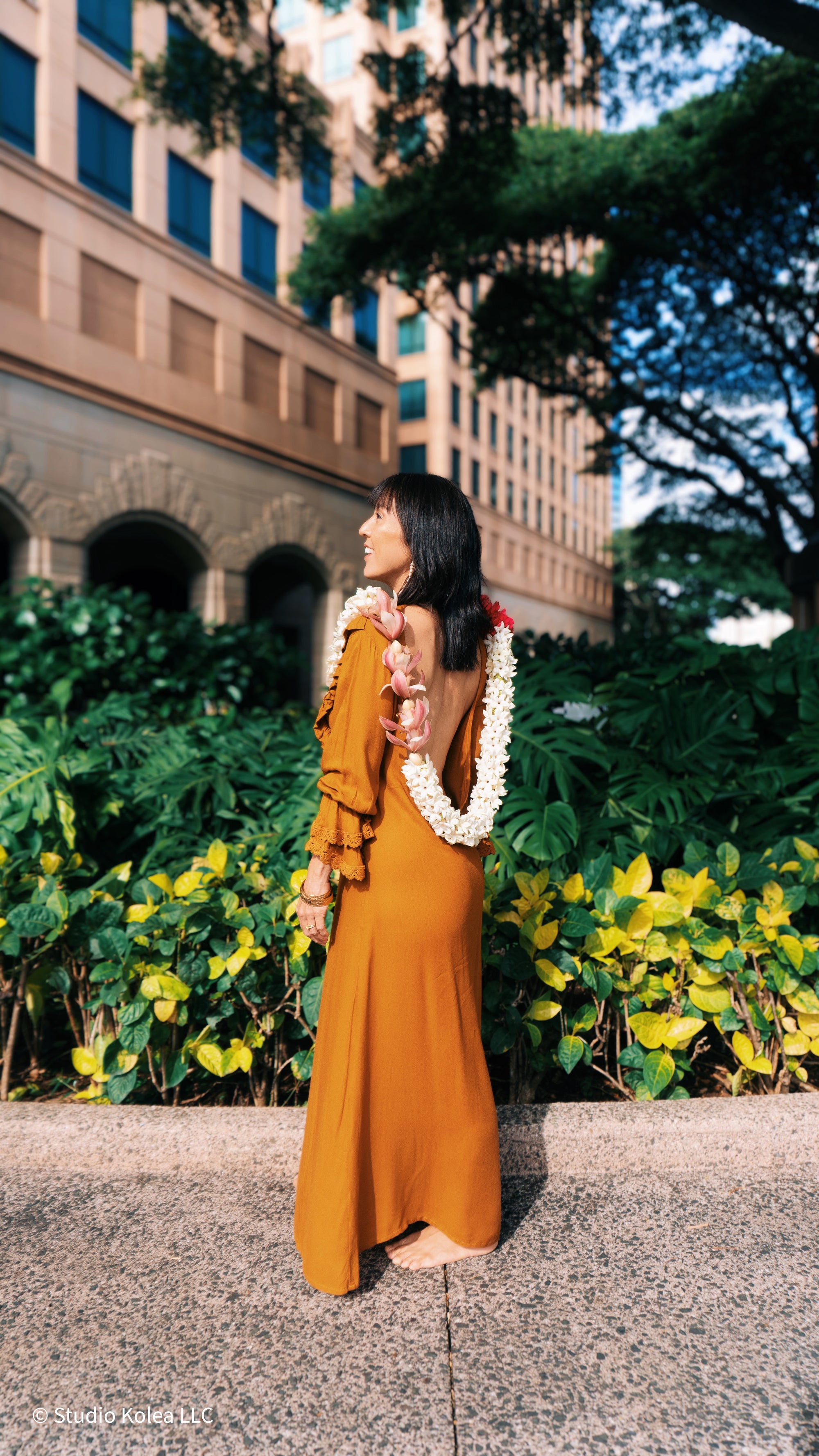 Woman in a yellow dress muumuu with a lei standing in front of a building with greenery.